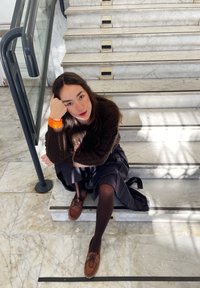 Persona seduta su scale di marmo, indossando un maglione di lana marrone, una gonna blu navy e scarpe marroni. Gli accessori includono un braccialetto arancione.