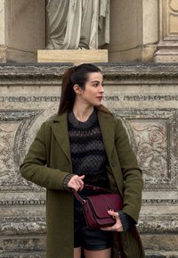 Femme en manteau vert et pull sombre tenant un sac à main bordeaux, debout devant un mur de pierre orné de détails sculptés et d'une statue.