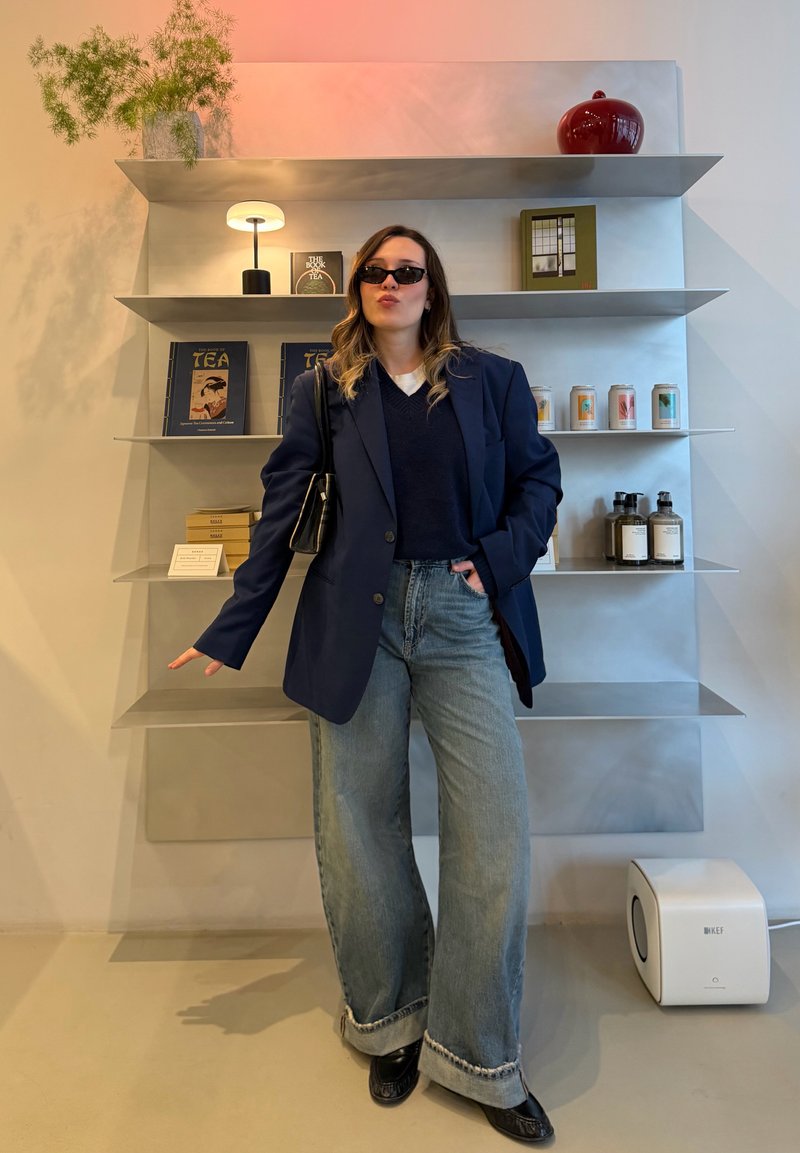 Femme portant des lunettes de soleil, un blazer foncé et un jean large, debout près d'étagères métalliques contenant des livres, des bougies et des lotions dans une pièce moderne et minimaliste.