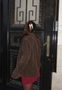 Braune Wildleder-Übergrößejacke, kurzer roter Rock, Haar mit Haarklammern gestylt. Im Hintergrund eine schwarze, kunstvoll gestaltete Tür mit satiniertem Glas.