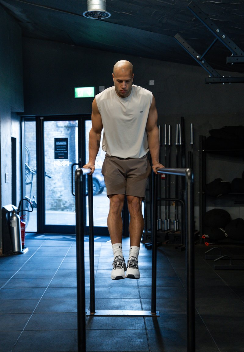 Homme en débardeur et short effectuant un exercice de dips aux barres parallèles dans une salle de sport avec du matériel de musculation en arrière-plan.
