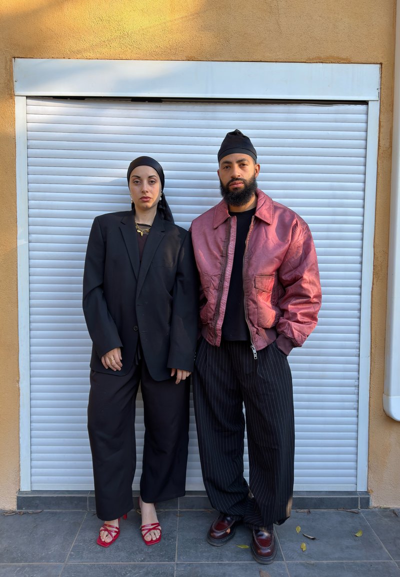 Deux personnes se tiennent côte à côte. La femme porte un blazer noir oversize et un pantalon à jambes larges, tandis que l'homme porte une veste bomber rose brillante et un pantalon à rayures.