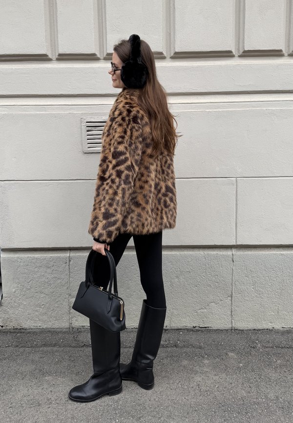 Femeie îmbrăcată într-o haină de blană artificială cu imprimeu de leopard, colanți negri, cizme până la genunchi, căști negre pentru urechi, ochelari, ținând o geantă de mână neagră, mergând lângă un perete palid.