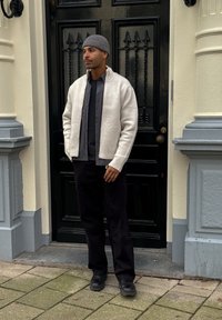 Cardigan tricoté gris clair porté sur une chemise noire, pantalon noir et bottes noires, avec un bonnet gris. Debout devant une porte noire.