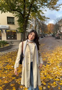 Nuori nainen beigenvärisessä takissa ja huivissa seisoo lehtien peittämällä kadulla, taustalla syksyisiä puita ja polkupyöriä.