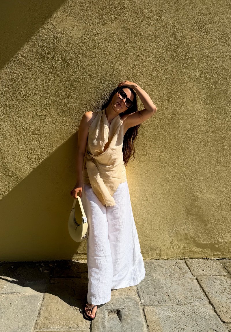 Femme portant des lunettes de soleil, un haut beige sans manches et un pantalon blanc à jambes larges, se tient contre un mur jaune texturé, tenant un sac à main clair et se touchant la tête.