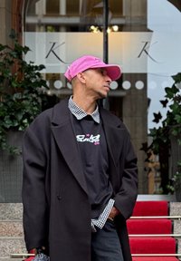 Melna jaka virs melna grafiskā T-kreklu ar rozā burtiem, redzama šaha raksta krekla apkakle, rozā cepure. Sarkanā paklāja un lapotnes fons.