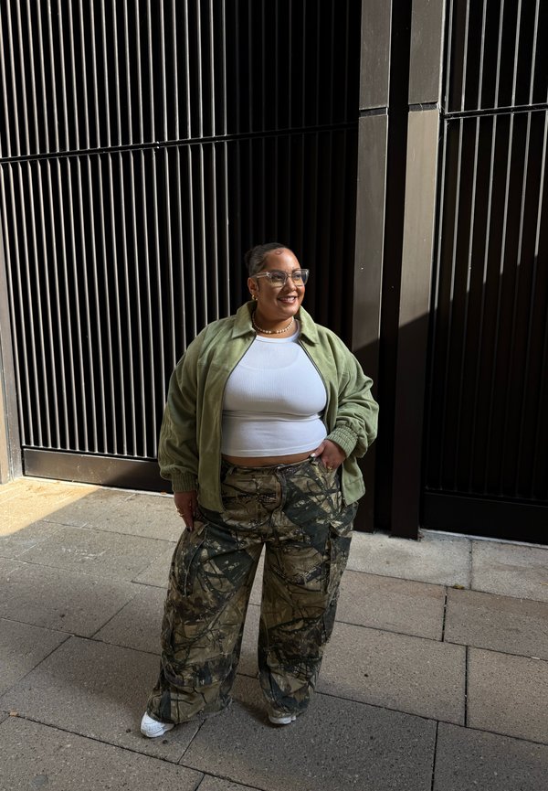Jachetă bomber verde măsliniu peste un crop top alb, pantaloni cargo cu model în nuanțe de verde și maro, adidași albi, stând la soare.