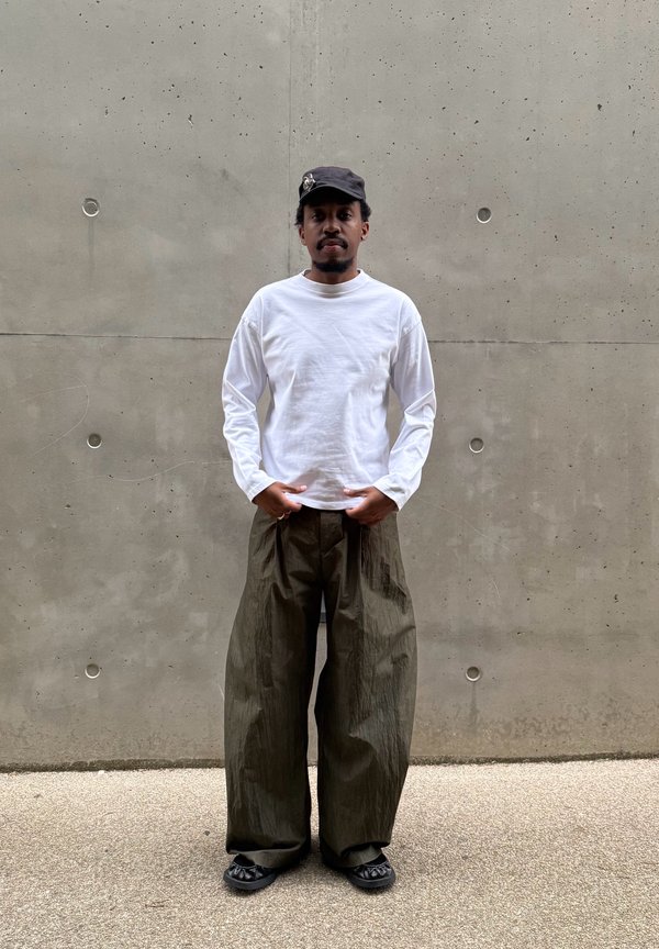 Homme portant une casquette noire, une chemise blanche à manches longues, un pantalon large kaki foncé et des chaussures noires, debout contre un mur en béton.