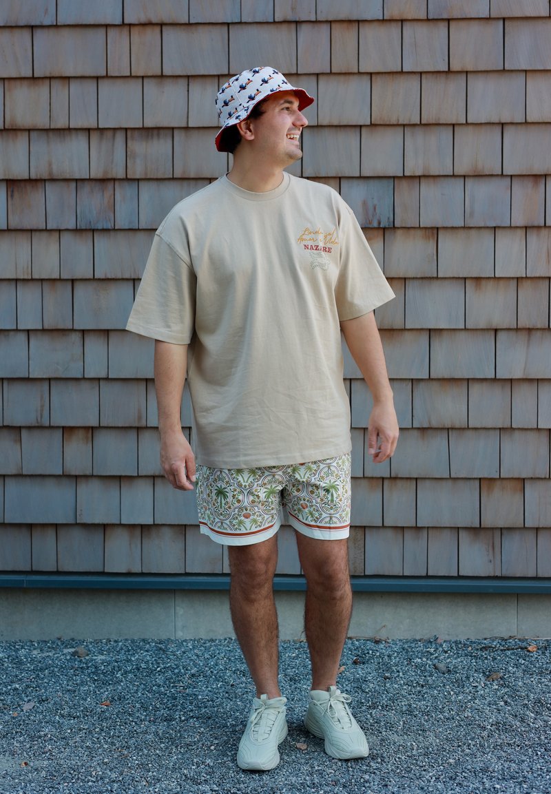 Homme portant un t-shirt beige oversize, un short à motifs, des baskets blanches et un bob blanc avec de petits motifs rouges et noirs, debout sur du gravier.