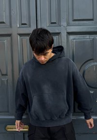 Νεαρός άνδρας με σκούρο φούτερ με κουκούλα που κοιτάζει προς τα κάτω, στέκεται μπροστά σε μια μεγάλη γκρι πόρτα με πάν ελ, με ένα χάλκινο θυρίδα αλληλογραφίας στο ύψος της μέσης.