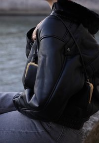 Schwarze Lederjacke mit einem mit Fell gefütterten Kragen, ausgestattet mit einem Reißverschluss und auffälligen Schulternähten. Kombiniert mit einer kleinen schwarzen Tasche und grauen Jeans.