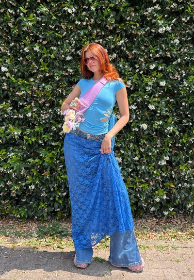 Top ajustado azul claro con cortes, superpuesto con una falda de encaje azul sobre jeans de campana, complementado con un bolso de tirante púrpura y acentos florales.