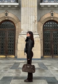 Kobieta w czarnej skórzanej kurtce, ciemnych spodniach i okularach przeciwsłonecznych stoi bokiem, trzymając dużą brązową torebkę z zamszu przed ozdobnymi drewnianymi drzwiami.