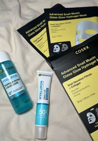 Sylinderformet transparent flaske med Garnier væske, to pakker med hvite og svarte COSRX hydrogel masker, og en hvit tube med Garnier solkrem.