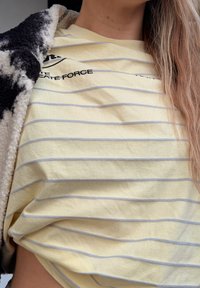 Άτομο που φοράει κίτρινη και λευκή ριγέ μπλούζα με μαύρο κείμενο, εν μέρει καλυμμένο από μαύρο και άσπρο χνουδωτό σακάκι, με ορατά μακριά ανοιχτό καστανά μαλλιά.