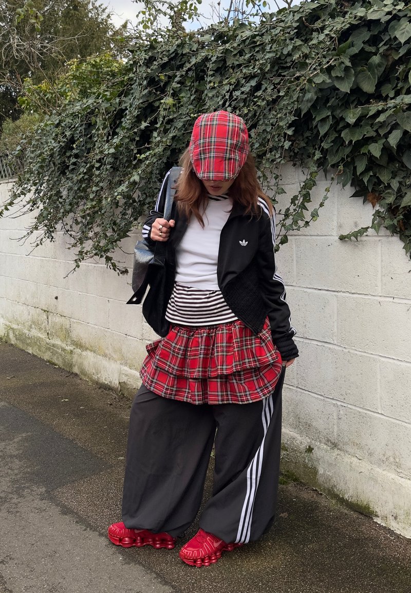 Personne portant une casquette à carreaux rouges et une jupe superposée sur un pantalon de survêtement noir Adidas et une veste, debout près d'un mur blanc avec du lierre vert.