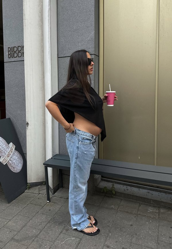 Femme enceinte avec des lunettes de soleil, portant un haut noir et un jean ample, tenant un gobelet rose avec une paille, debout à côté d'un banc sur un trottoir en ville.