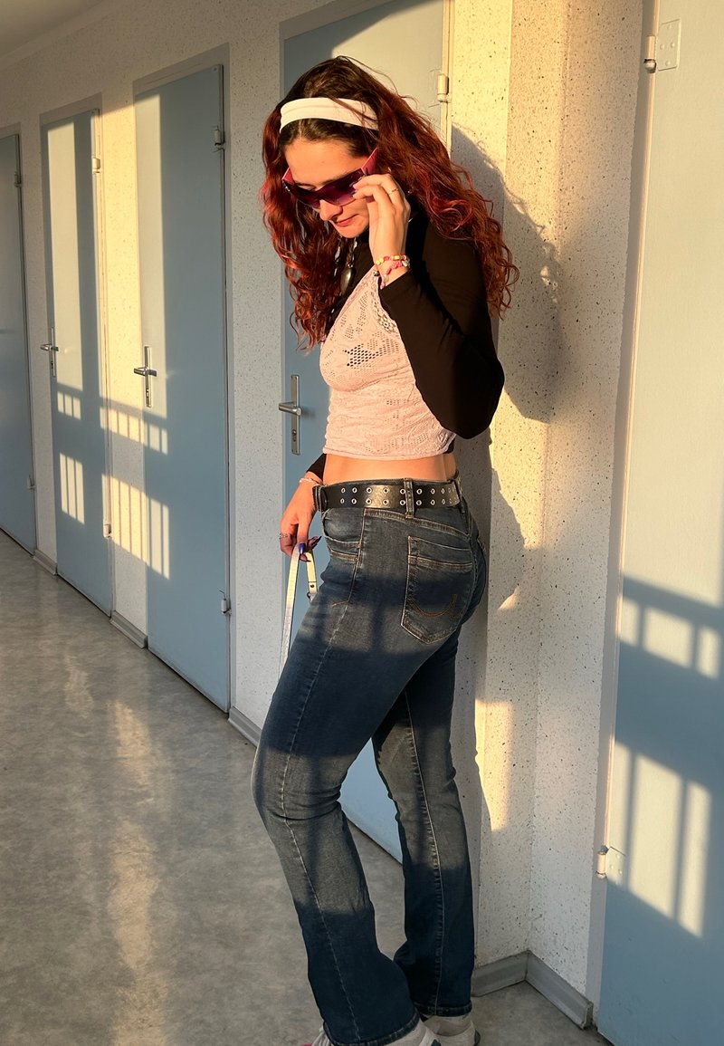Lyserød blonde crop top over en sort langærmet skjorte, parret med mørkeblå flared jeans og hvide sneakers, accessorized med solbriller.