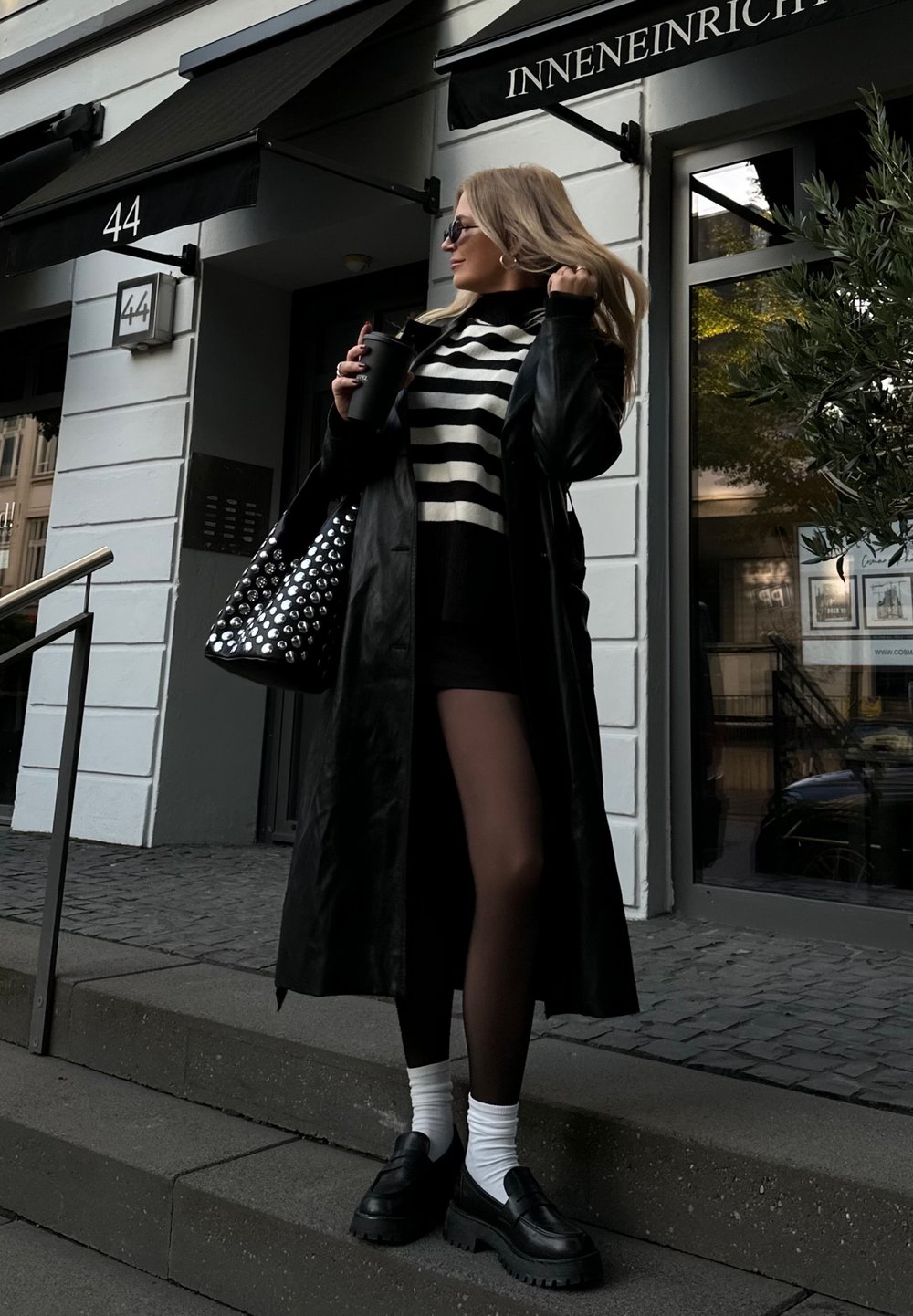 Μαύρο δερμάτινο μακρύ παλτό, μαύρο και λευκό ριγέ πουλόβερ, κοντή φούστα, μαύρες πλατφόρμες loafers και τσάντα με καρφιά. Υπαίθριο περιβάλλον.