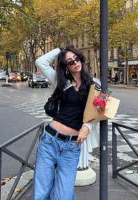 Jonge vrouw met een zonnebril houdt een boeket vast, draagt een zwart topje en blauwe jeans, en staat op een stadsstraat met herfstbomen en verkeer op de achtergrond.