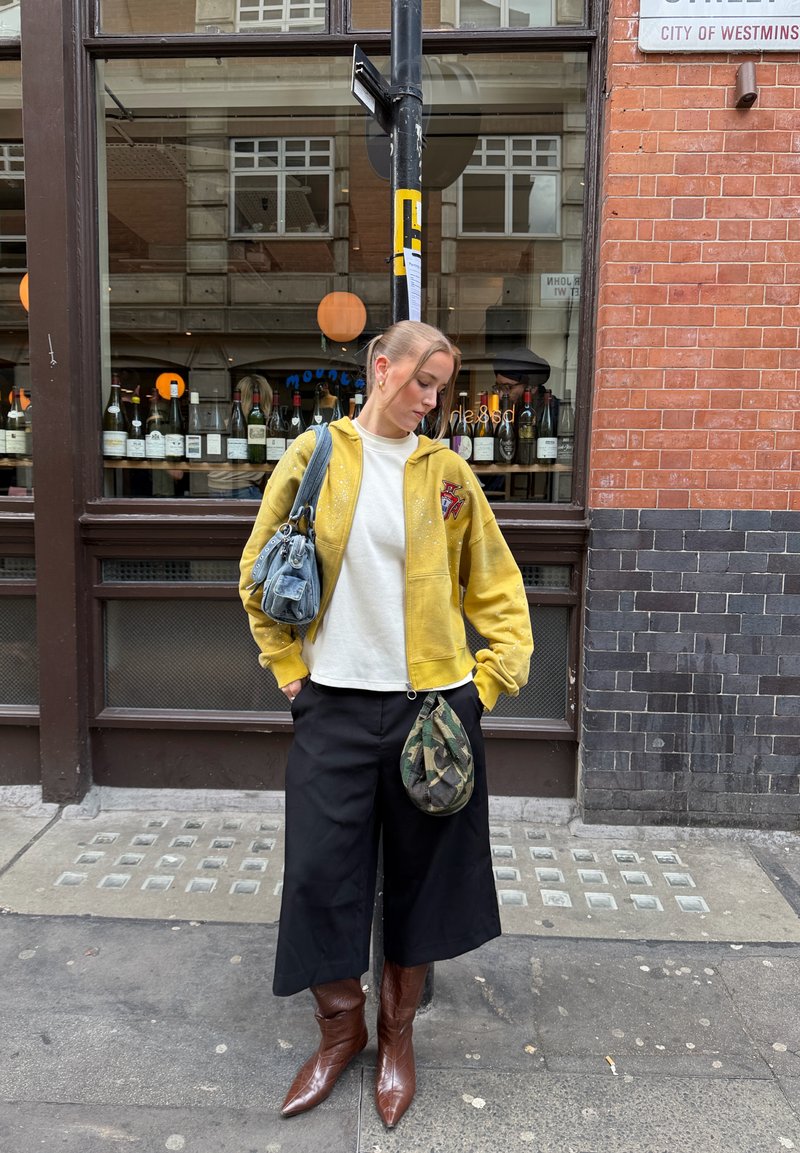 Jeune femme en veste jaune et pantalon large noir se tient devant une fenêtre avec des bouteilles de vin, porte des bottes marron et porte deux sacs.