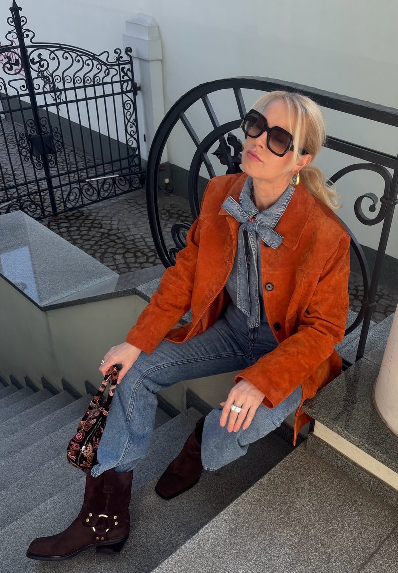 Frau trägt orange Wildlederjacke, Schleifenbluse aus Denim, Jeans, dunkle Stiefel und Sonnenbrille, sitzt auf Steinstufen und hält eine gemusterte Handtasche.