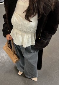 Vluchtige witte peplumblouse, donkere nepbonten jas, grijze broek met wijde pijpen, tan suède handtas, en naakte schoenen op een betonnen vloer.