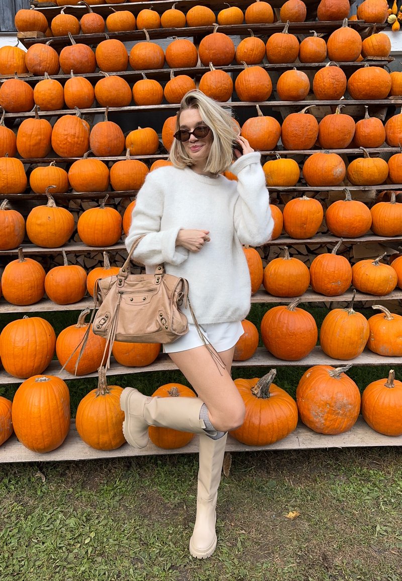 Femme en pull blanc, shorts, bottes beiges et lunettes de soleil posant devant un support en bois avec plusieurs citrouilles orange.