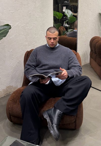 Sillón de tela marrón con un diseño redondeado. Persona sentada, vistiendo un suéter gris sobre una camisa clara, leyendo una revista. Botas negras visibles.