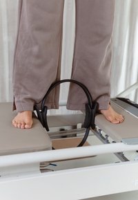 Pilates reformer med en beige polstret plattform og svart motstandsring. Bare føtter plassert på utstyret; myke stoffbukser synlige.