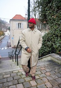 Άνδρας με μπεζ παλτό, κόκκινο σκουφάκι, γυαλιά και καφέ παπούτσια που στέκεται σε λιθόστρωτα σκαλιά κοντά σε φυλλώματα και σε δρόμο με κτίρια στο βάθος.