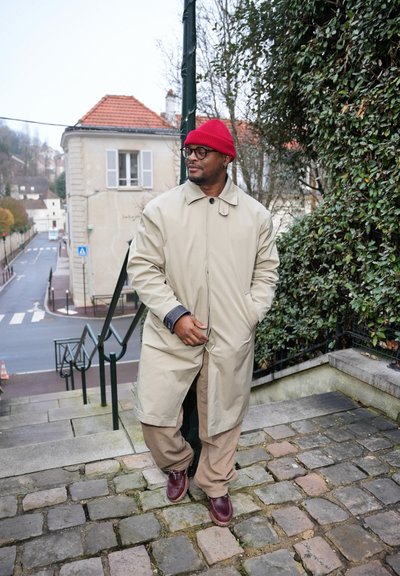 Hombre con abrigo beige, gorro rojo, gafas y zapatos marrones de pie en escalones de adoquines cerca de vegetación y una calle con edificios al fondo.