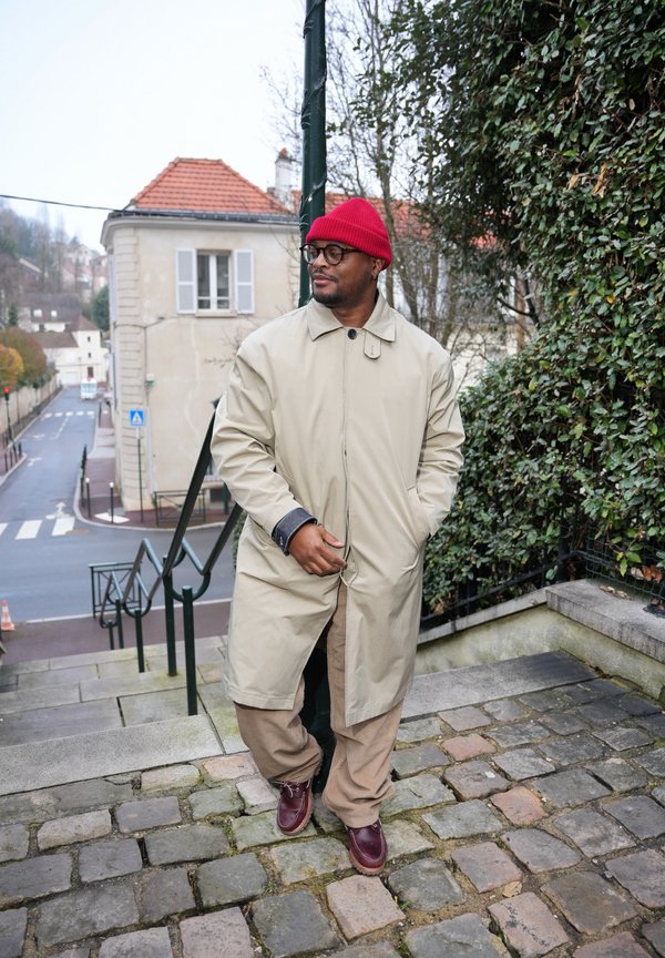 Mann iført beige kåpe, rød lue, briller og brune sko som står på brosteinsbelagte trinn nær grøntområder og en gate med bygninger i bakgrunnen.