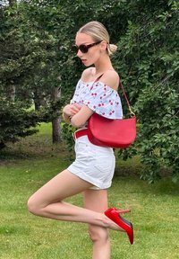 Borsa a mano rossa in pelle con design curvo e hardware dorato, indossata con un top a spalle scoperte a fantasia ciliegia e pantaloni corti bianchi. Tacchi alti rossi.