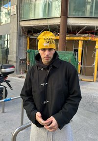 Čierna bunda so striebornými kovaniami, sivé tepláky a žltá čiapka s bielym textom. Textúry sú hladké s neformálnym dizajnom.