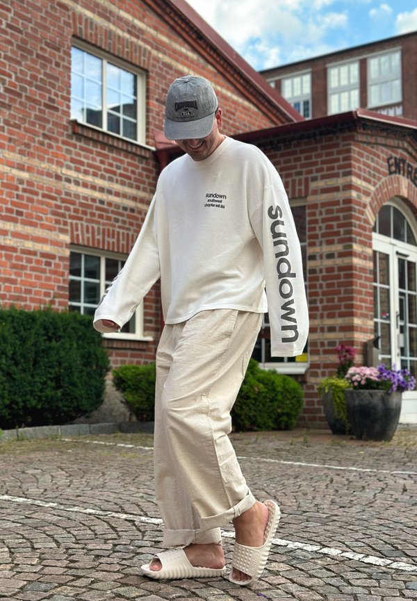Mann trägt beige Hose, weißes Langarmhemd mit dem Text "sundown", graue Kappe und weiße Slip-on-Sandalen, steht vor einem Ziegelgebäude.