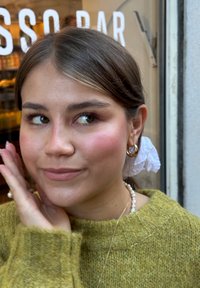 Gros plan sur une personne ayant les cheveux châtain clair, portant un pull en laine vert. Les traits incluent des boucles d'oreilles en or, un maquillage subtil et un élastique à cheveux blanc.