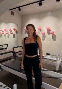 Sort atletisk top og bukser med en tætsiddende pasform. Baggrunden har lyserøde vægmonterede bolde og en hvid reformermaskine.