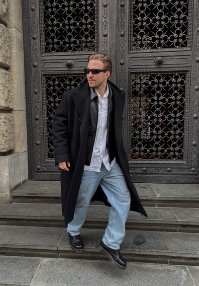 Largo abrigo negro sobre una camisa blanca y un chaleco negro, combinado con jeans de mezclilla azul claros y zapatos negros; de pie en unos escalones de piedra cerca de una puerta decorativa.