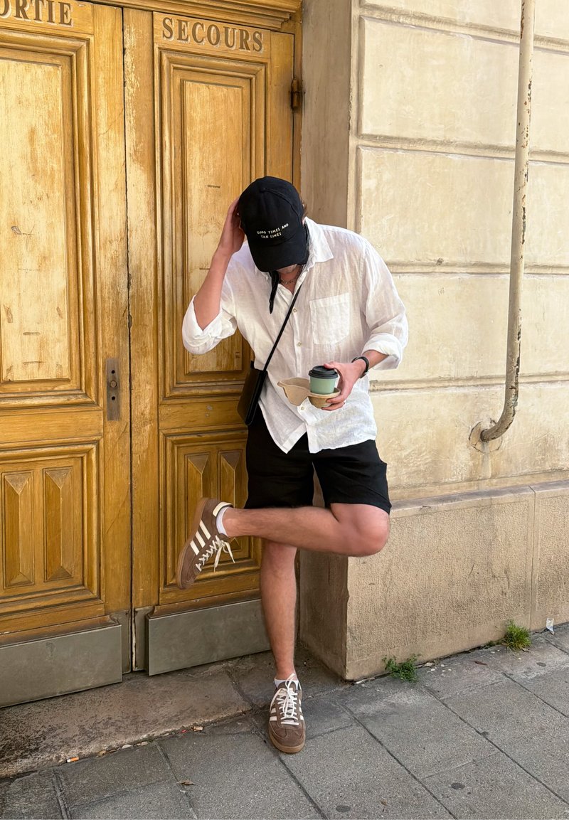 Person, die eine schwarze Kappe, ein weißes Hemd, schwarze Shorts und braune Turnschuhe trägt, steht auf einem Bein und hält eine Kaffeetasse neben einer Holztür mit der Aufschrift "Sortie Secours."