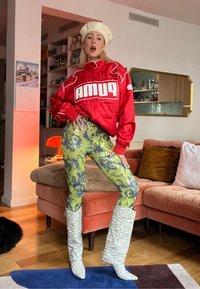 Femme posant à l'intérieur, portant un béret blanc, une veste rouge Puma, un pantalon jaune à motifs et de grandes bottes blanches texturées dans un salon.