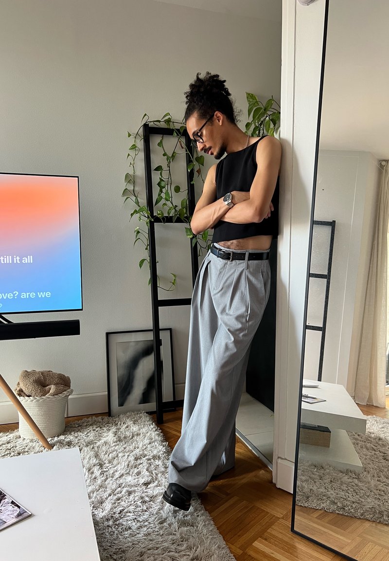 Personne avec des lunettes et des cheveux bouclés, portant un haut noir sans manches et un pantalon large gris, appuyée contre un mur près d'un grand miroir.