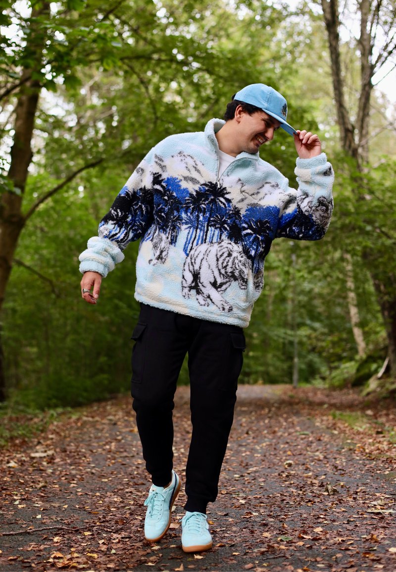 Pull en polaire avec base bleu clair, motifs de palmiers et de tigres noirs et bleus. Associé à un pantalon cargo noir et des baskets bleu clair.
