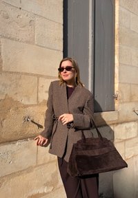 Brauner karierten Blazer mit Gürtel, dunkelbraune Wildleder-Handtasche und tiefviolettes Oberteil vor einer strukturierten Steinmauer.