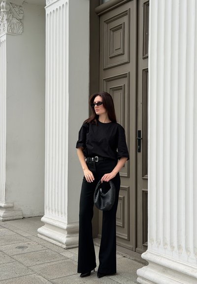 Mujer con conjunto negro y gafas de sol sosteniendo un bolso negro, de pie entre altas columnas blancas cerca de una puerta de madera ornamentada.
