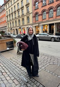 Manteau noir, écharpe grise et pull noir surdimensionné porté avec un pantalon large. Tenant un sac à main tressé bordeaux foncé, debout sur une rue pavée.