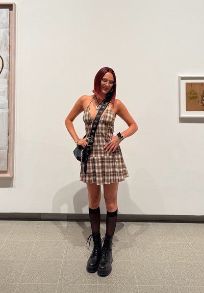 Vestido a cuadros marrón con dobladillo acampanado, medias altas negras hasta la rodilla y botas gruesas negras. Los accesorios incluyen un bolso cruzado y joyas en capas.