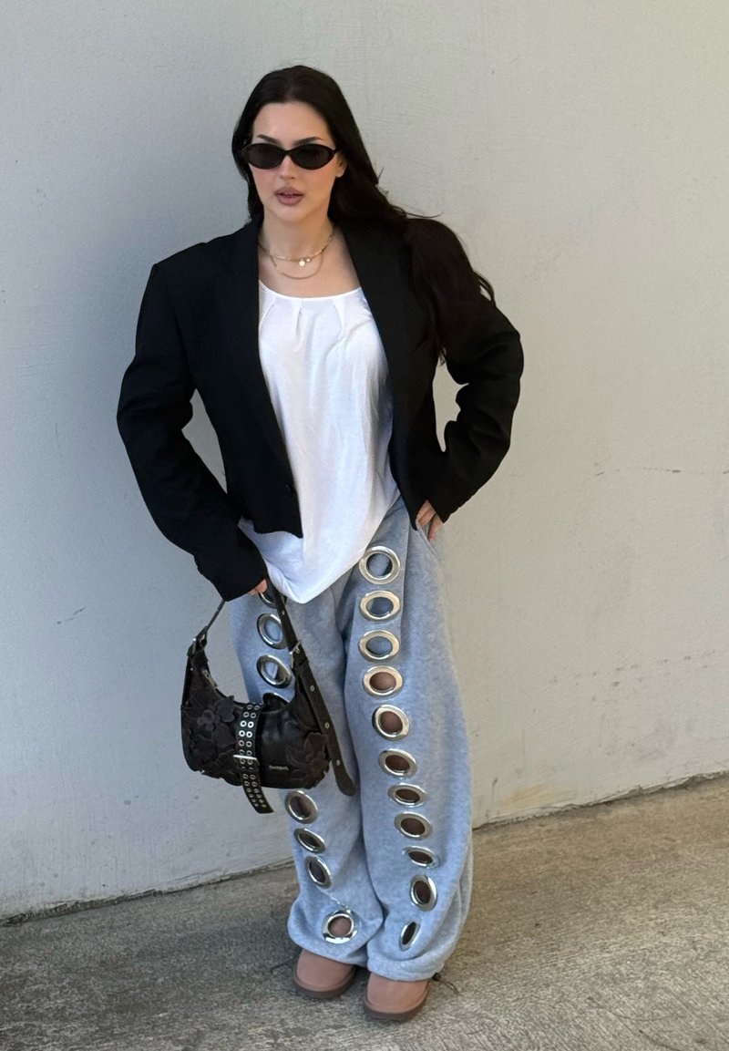 Femme en blazer noir surdimensionné, haut blanc, pantalon gris avec grands œillets argentés, lunettes de soleil, tenant un sac à main noir, debout contre un mur uni.