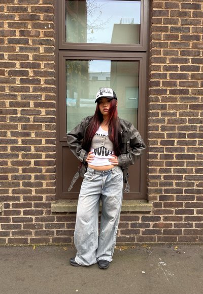 Chaqueta de cuero negra, top corto blanco con estampado, pantalones vaqueros holgados de color azul claro y una gorra en blanco y negro. Fondo: pared de ladrillo y ventana.